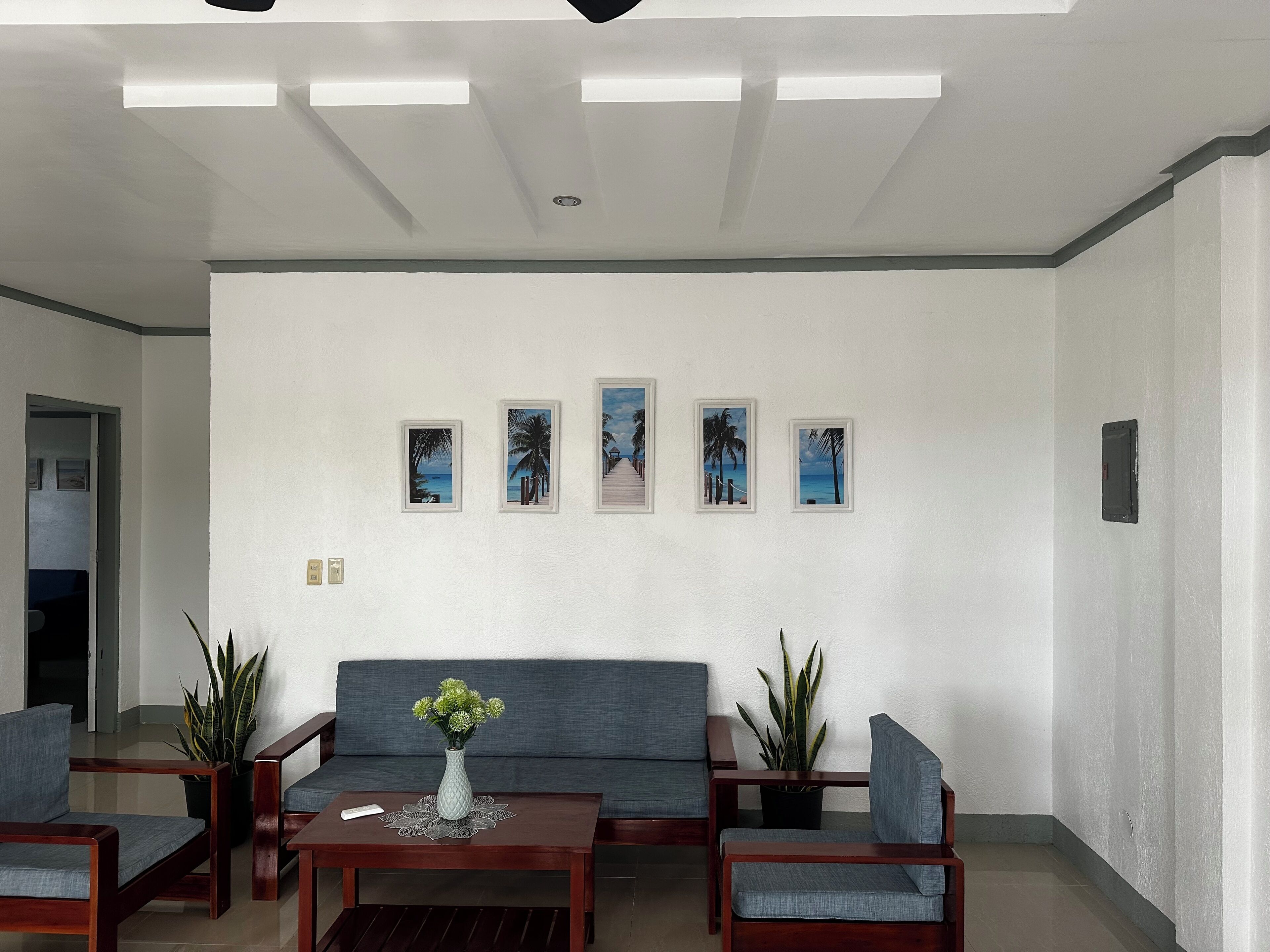 A modern living room with a grey sofa, wooden coffee table, and two armchairs. There are five framed pictures on the wall depicting tropical scenes.