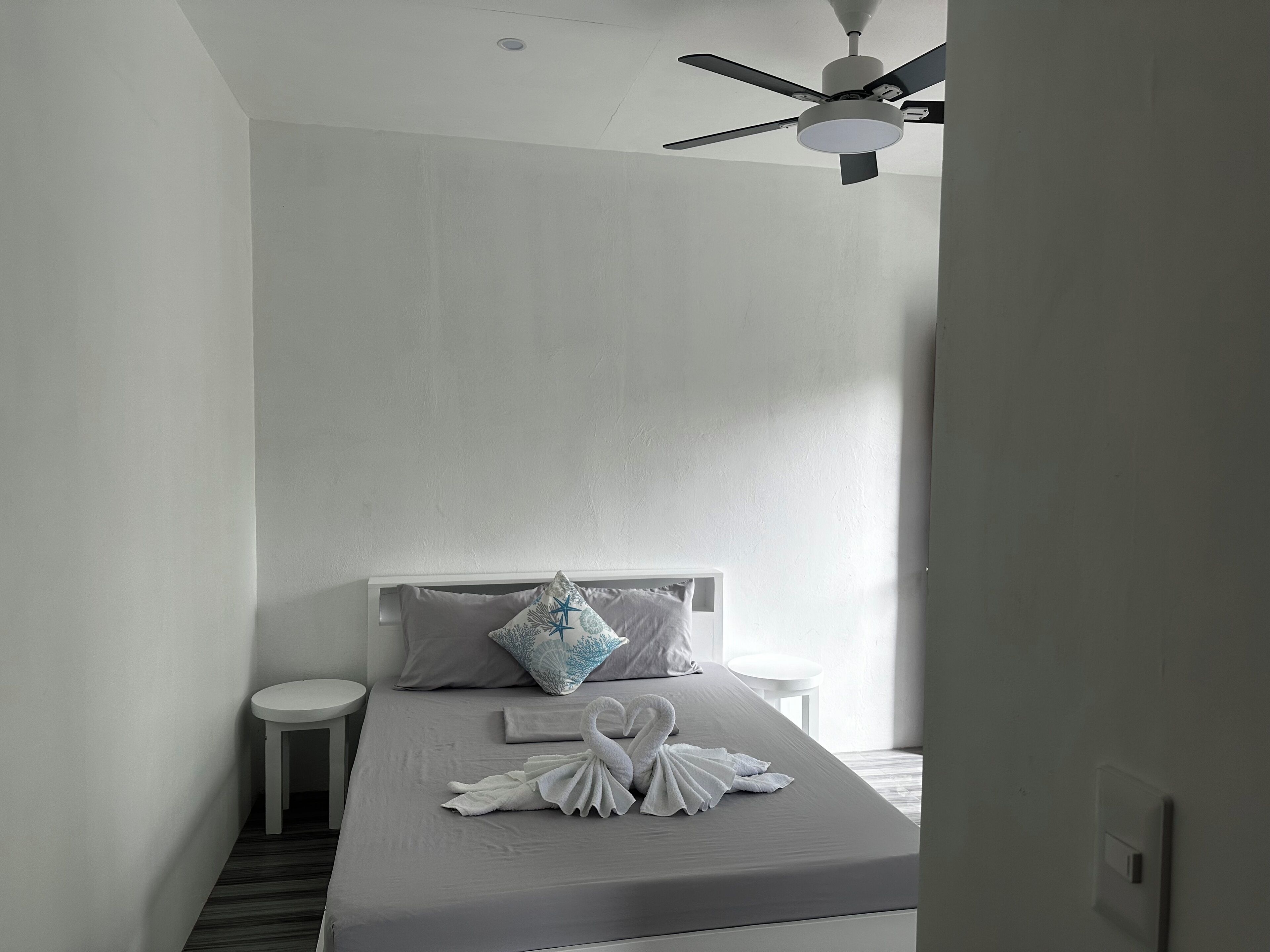 A minimalist hotel room with a bed, a small bedside table, a ceiling fan, and a window allowing natural light.