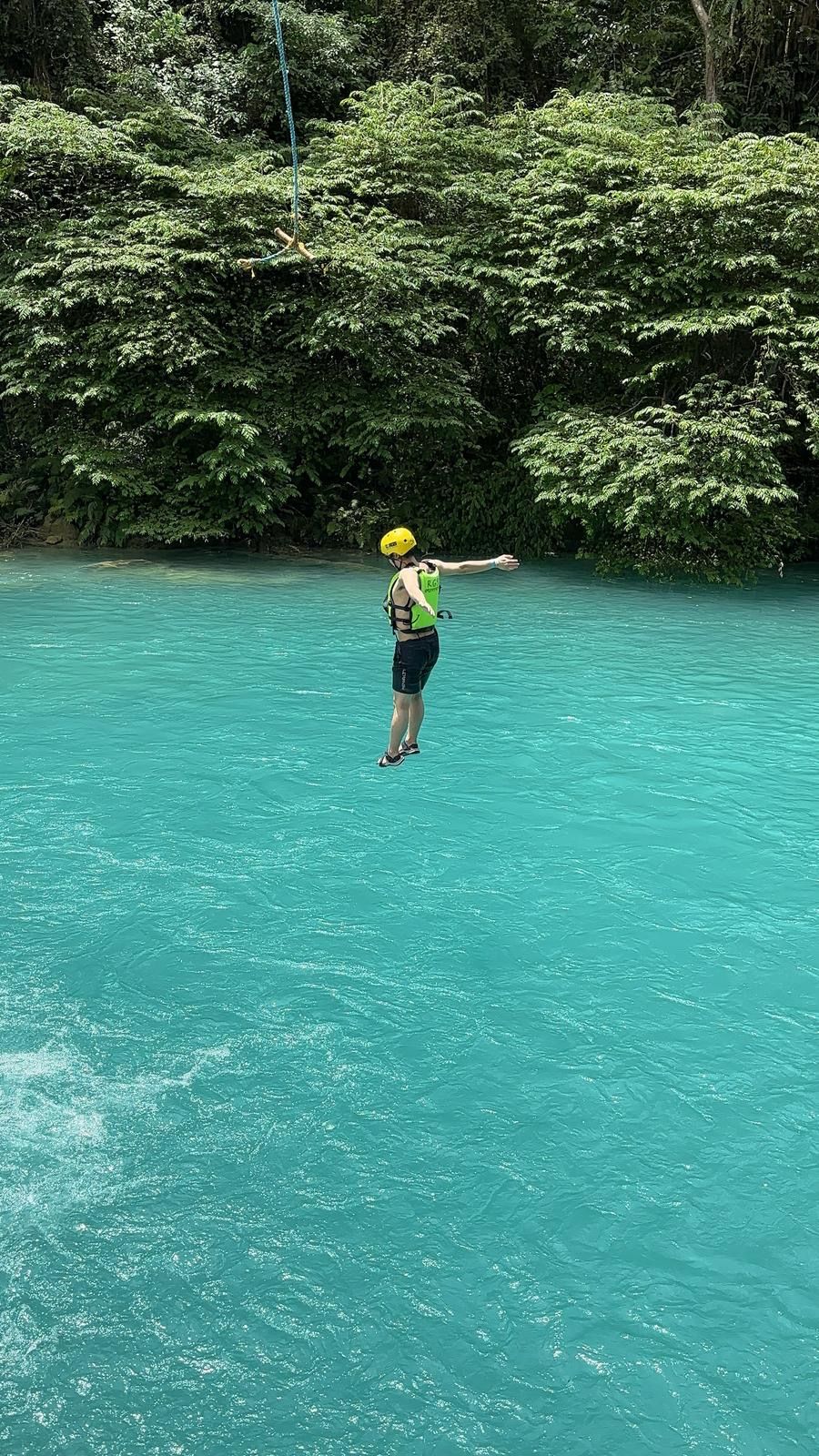 A person in a green life jacket and yellow helmet standing on a floating platform in a turquoise body of water surrounded by dense green foliage.