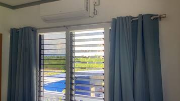 A room with a window featuring horizontal blinds, a ceiling-mounted air conditioning unit, and a curtain rod.