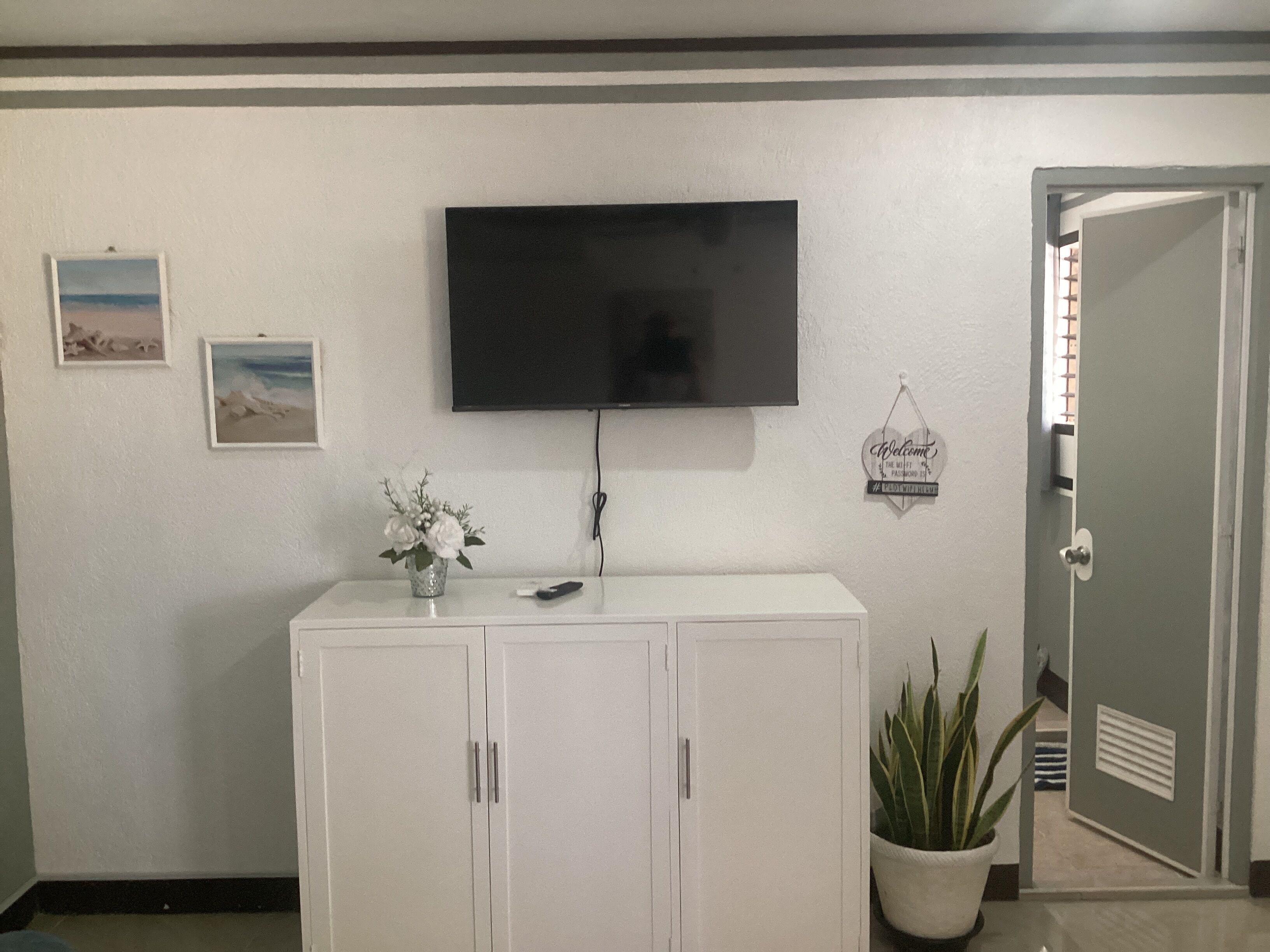 A room with a flat-screen TV mounted on the wall, a white cabinet with two doors, a potted plant, and a welcome sign.