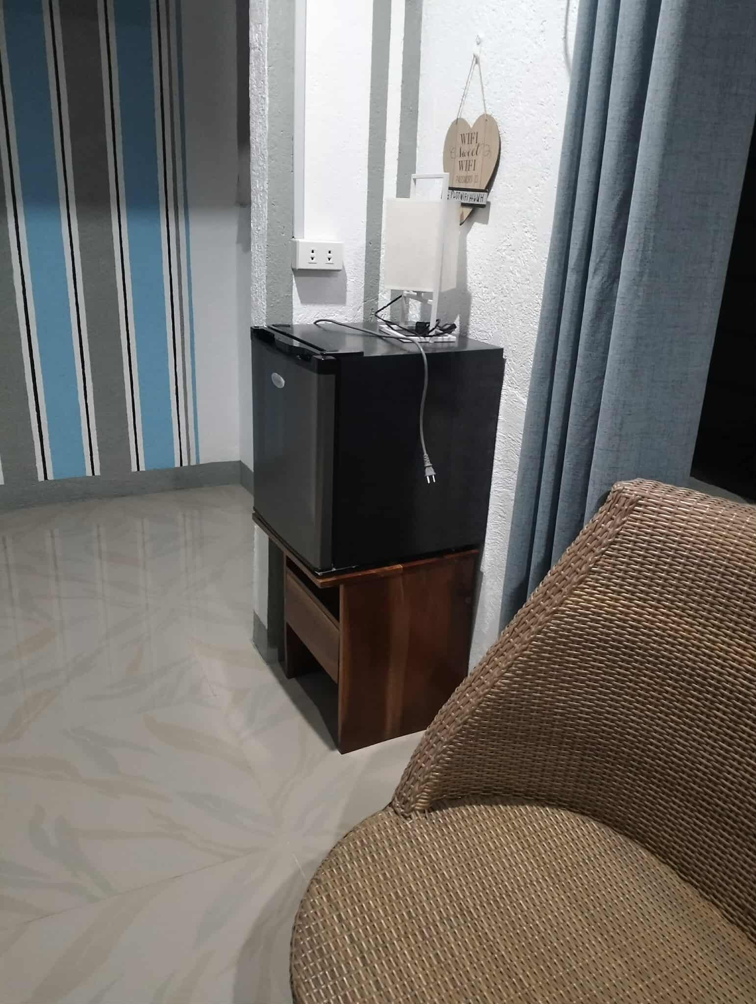 Quarto compacto com uma pequena TV preta em um suporte de madeira, uma cadeira de vime e um letreiro fixado na parede indicando a disponibilidade de WiFi.