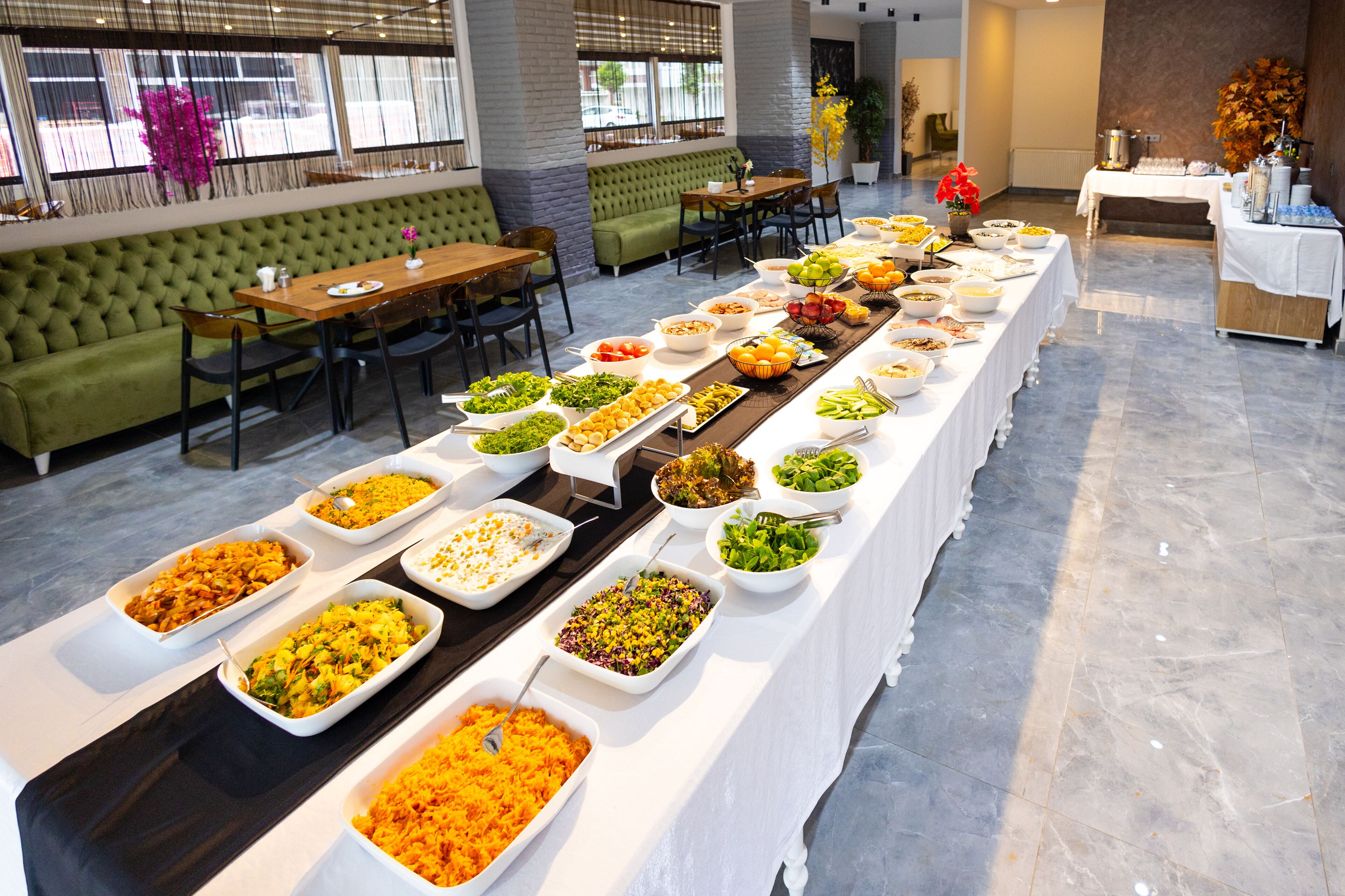 Montaje de bufé con diversos alimentos dispuestos en una mesa larga, rodeado de asientos y un sofá verde.
