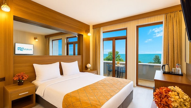 A hotel room with a large bed, a nightstand, a desk, a chair, and a balcony with a view of the sea.
