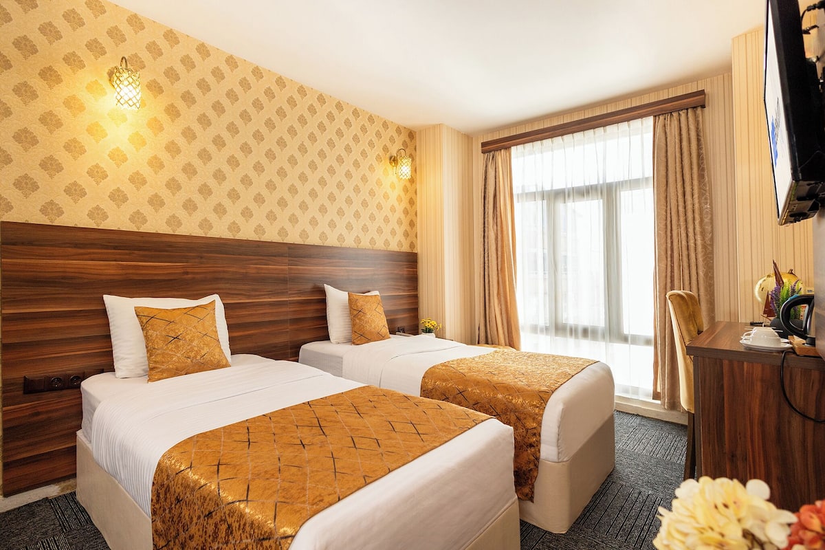 Ein Hotelzimmer mit zwei Betten, einem Holz-Kopfteil, einem Schreibtisch mit Stuhl, einem Fernseher und einem Fenster mit Vorhängen.