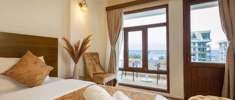 A hotel room with a bed, a view of the sea, a balcony with outdoor seating, and a wooden headboard.