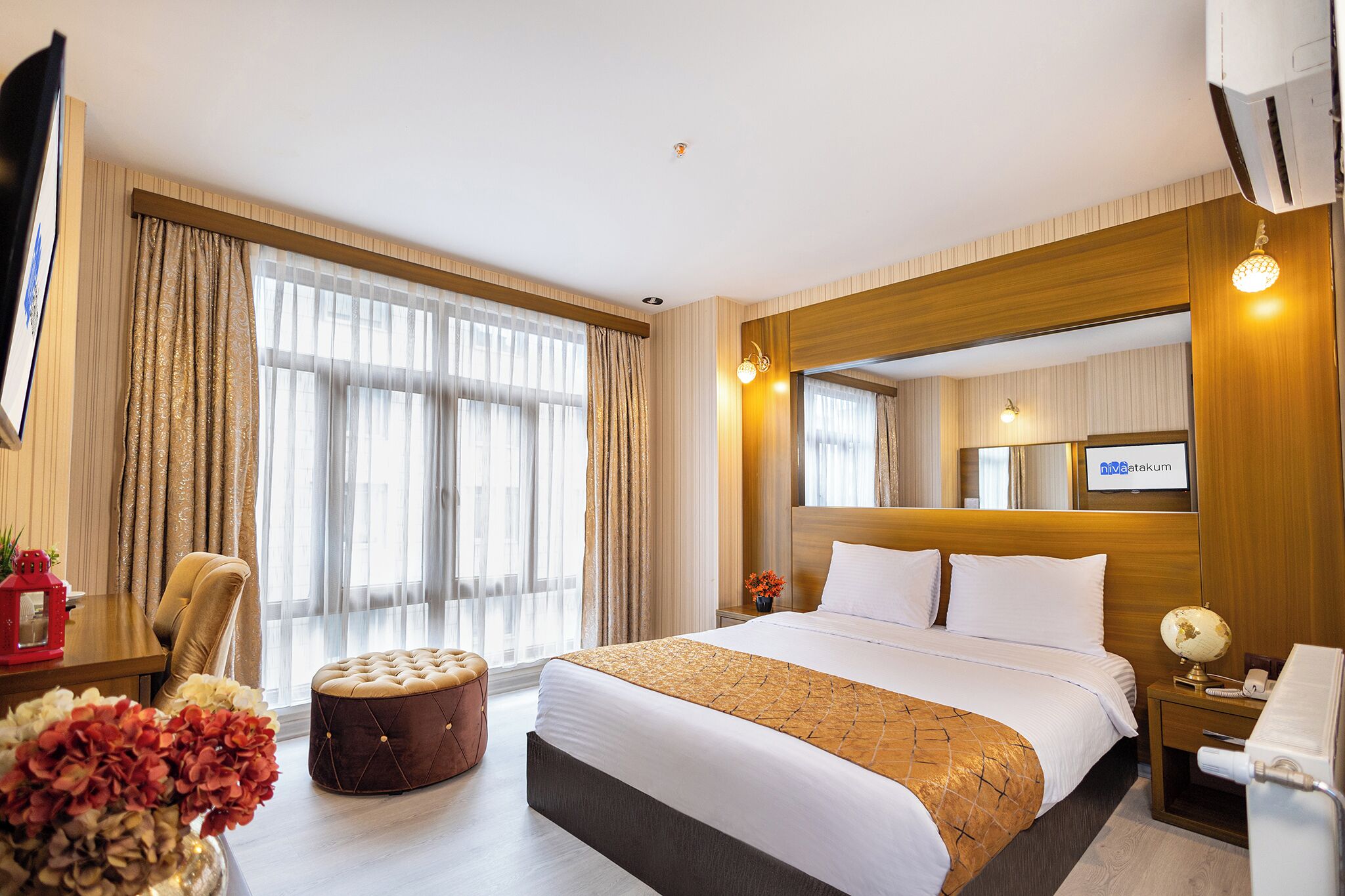 Una habitación de hotel con una cama grande, un cabecero de madera, un televisor, una mesita de noche con una lámpara y una ventana con cortinas.
