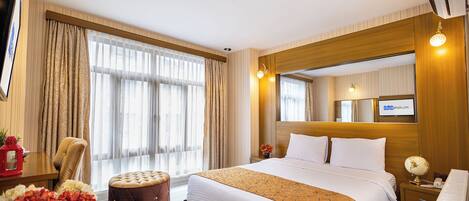 A hotel room with a large bed, a wooden headboard, a TV, a bedside table with a lamp, and a window with curtains.