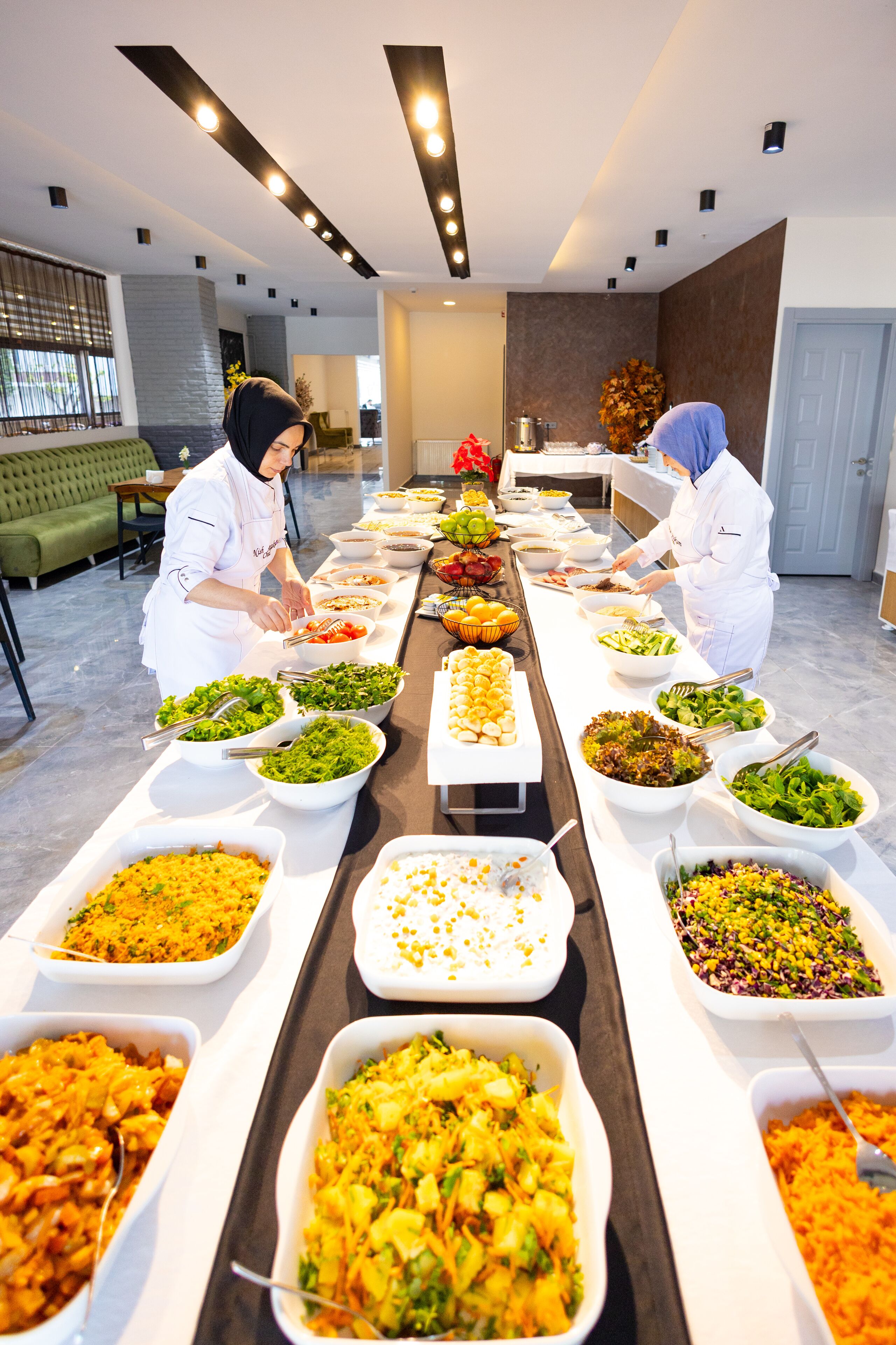 A buffet setup with various dishes in white bowls and trays, arranged on a long table in a well-lit room with a green sofa and a decorative plant in the background.