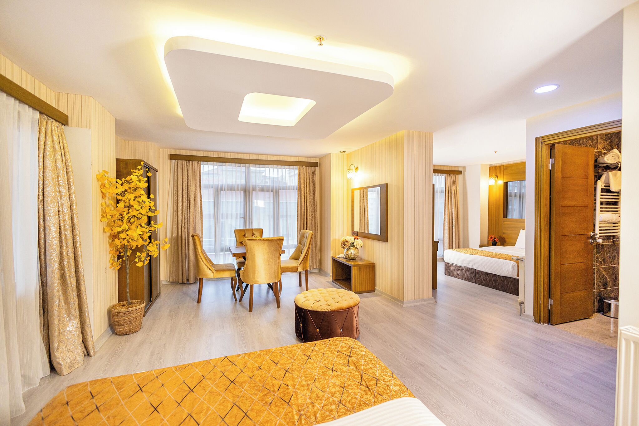 Quarto de hotel com cama, área de refeições com cadeiras, pequena mesa, planta em vaso e banheiro com pia e espelho.