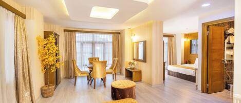 Quarto de hotel com cama, área de refeições com cadeiras, pequena mesa, planta em vaso e banheiro com pia e espelho.
