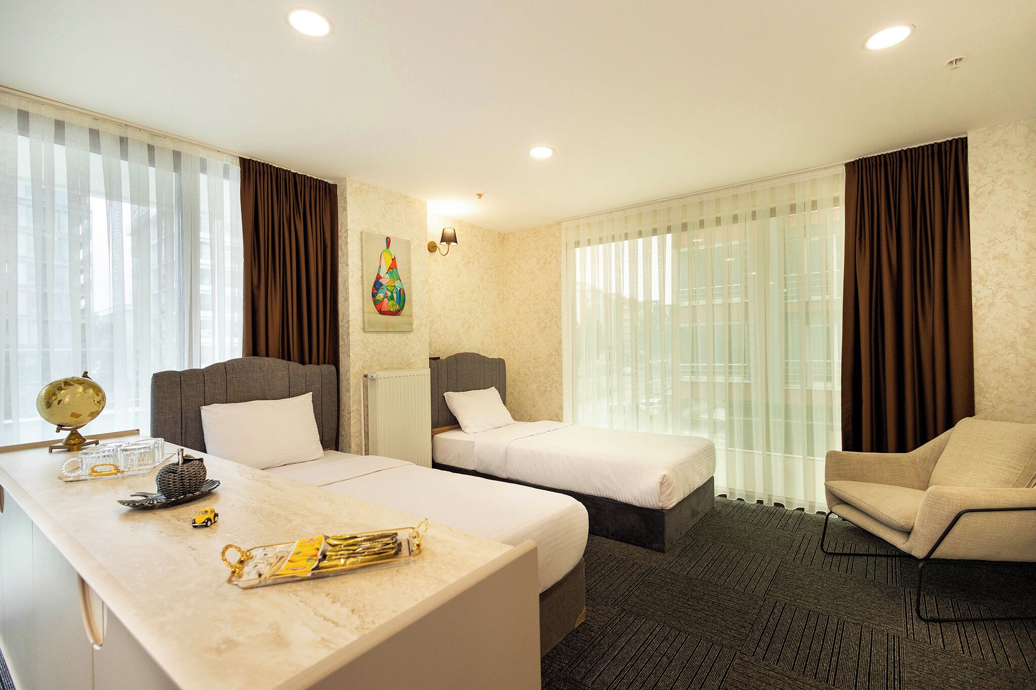 Habitación de hotel con dos camas, un escritorio con un globo terráqueo y una bandeja, una silla y un amplio ventanal con cortinas.
