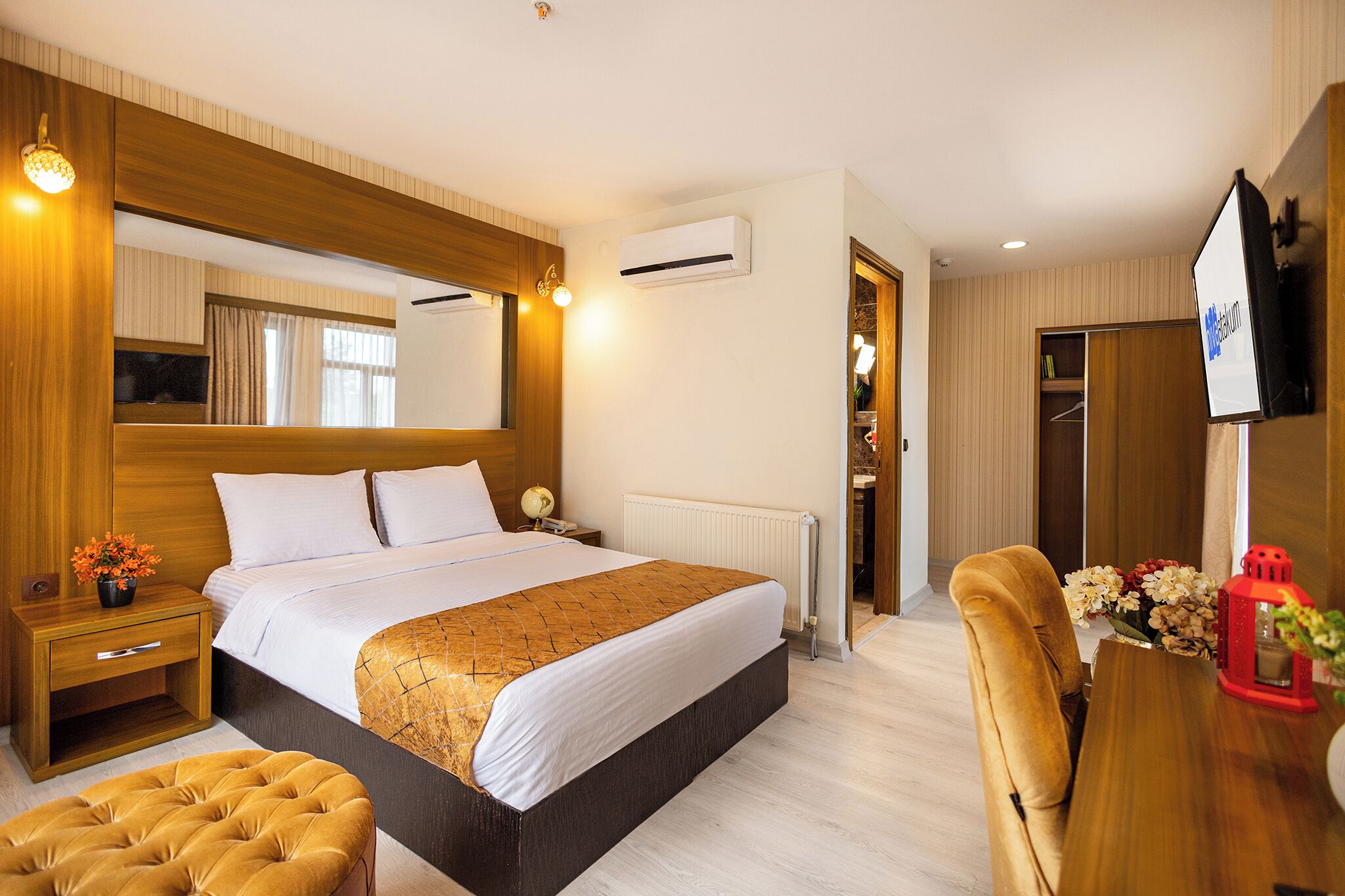 A hotel room with a large bed, wooden headboard, bedside table with a lamp, a small table with flowers, and a TV mounted on the wall.