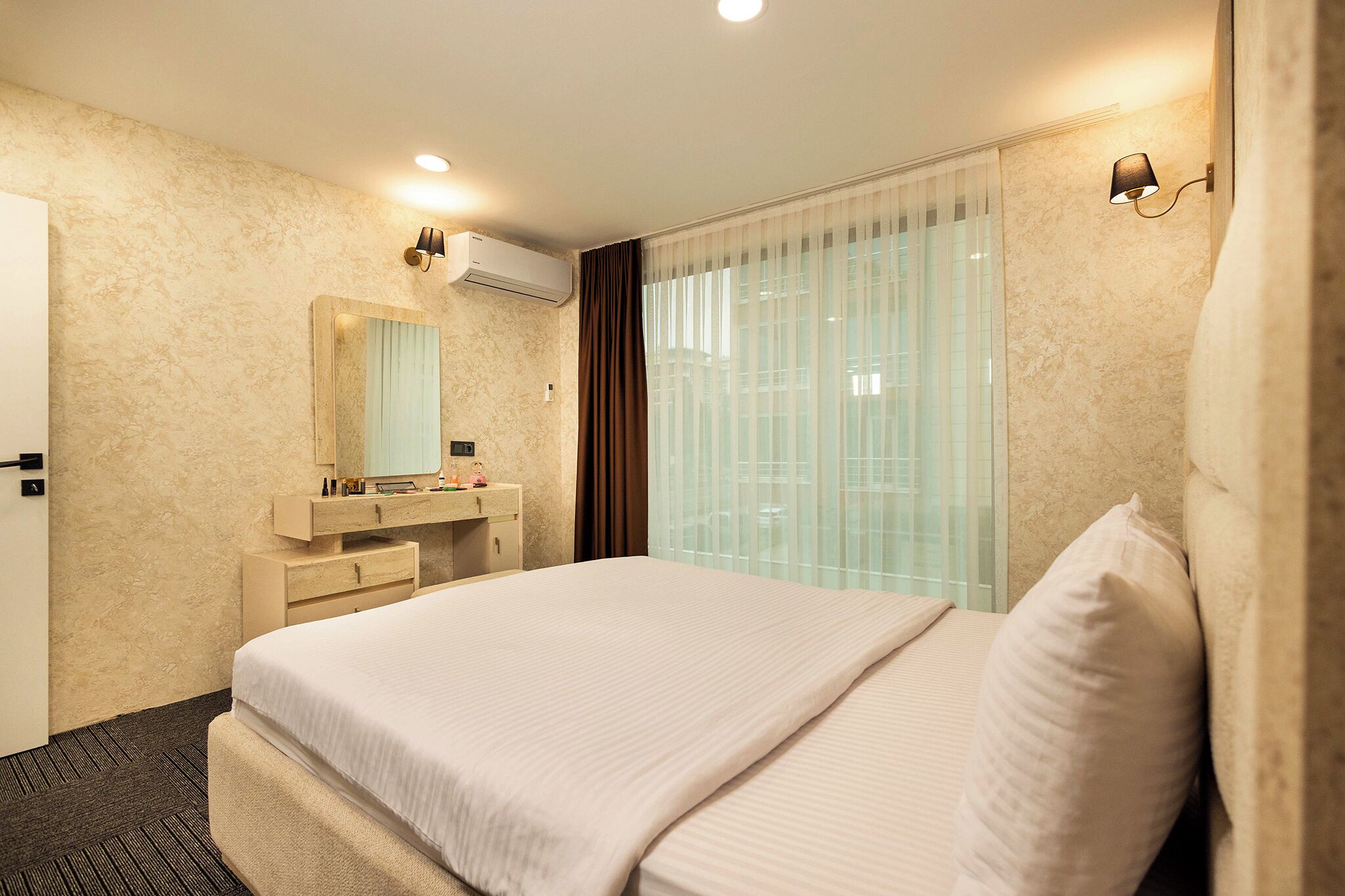 Une chambre d’hôtel avec un grand lit, une table de chevet avec un miroir, une petite commode et une fenêtre avec des rideaux légers.