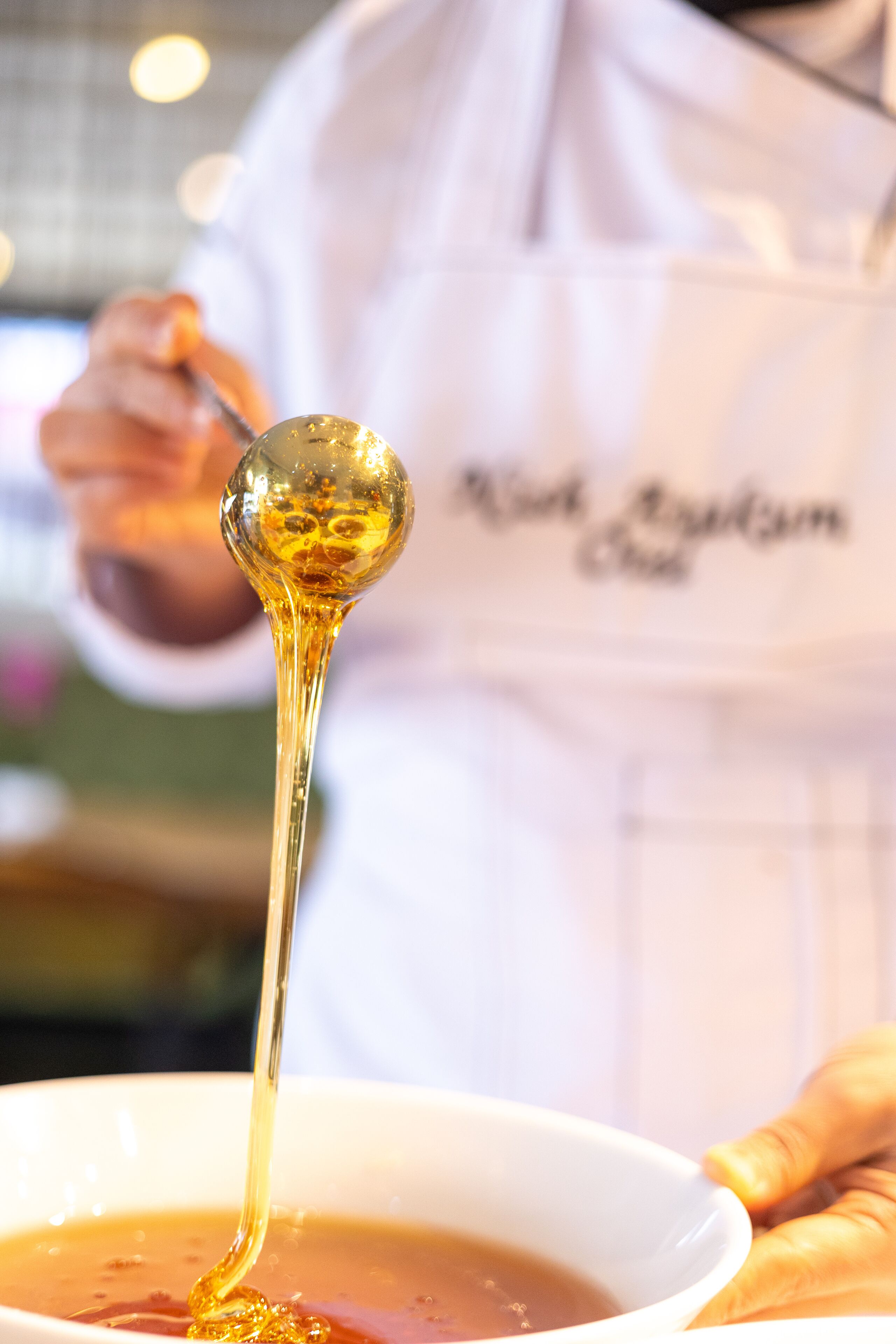 Una persona con bata de chef sosteniendo una cuchara grande con un líquido dorado y espeso.