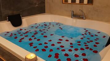 Una bañera llena de agua y pétalos de rosa, una vela encendida y un espejo con artículos de tocador.