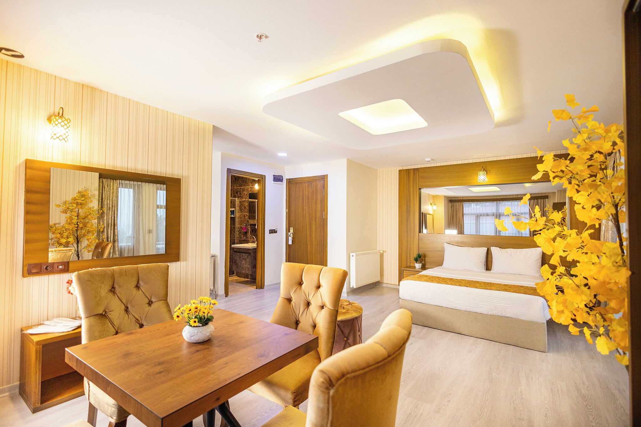 A modern hotel room with a wooden dining table, comfortable seating, a large bed, and a view of the outdoors.