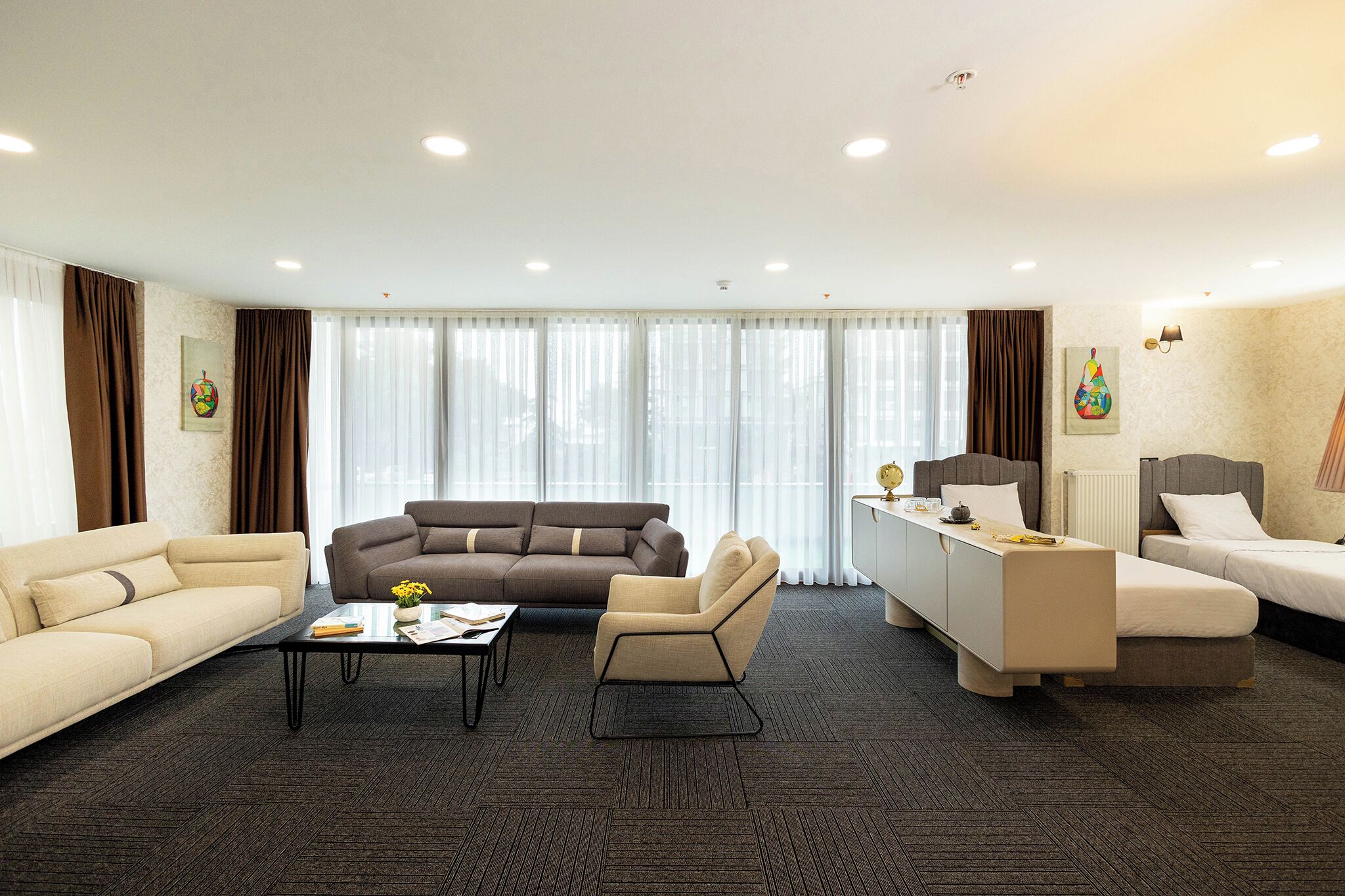 A hotel room with a sofa, armchair, a small table with a vase, a bed, and a desk with a lamp.