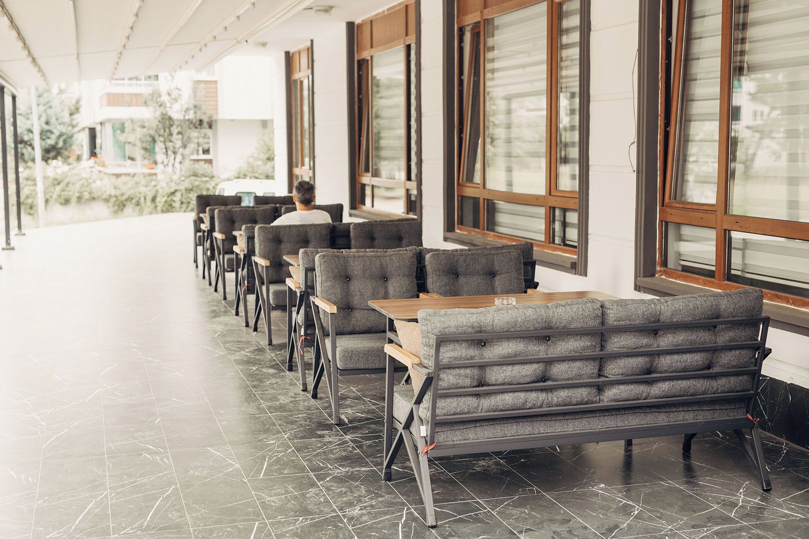 A row of grey upholstered seating with wooden tables, arranged in a linear fashion along a corridor with large windows.