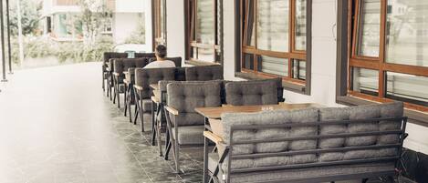 A row of grey upholstered seating with wooden tables, arranged in a linear fashion along a corridor with large windows.