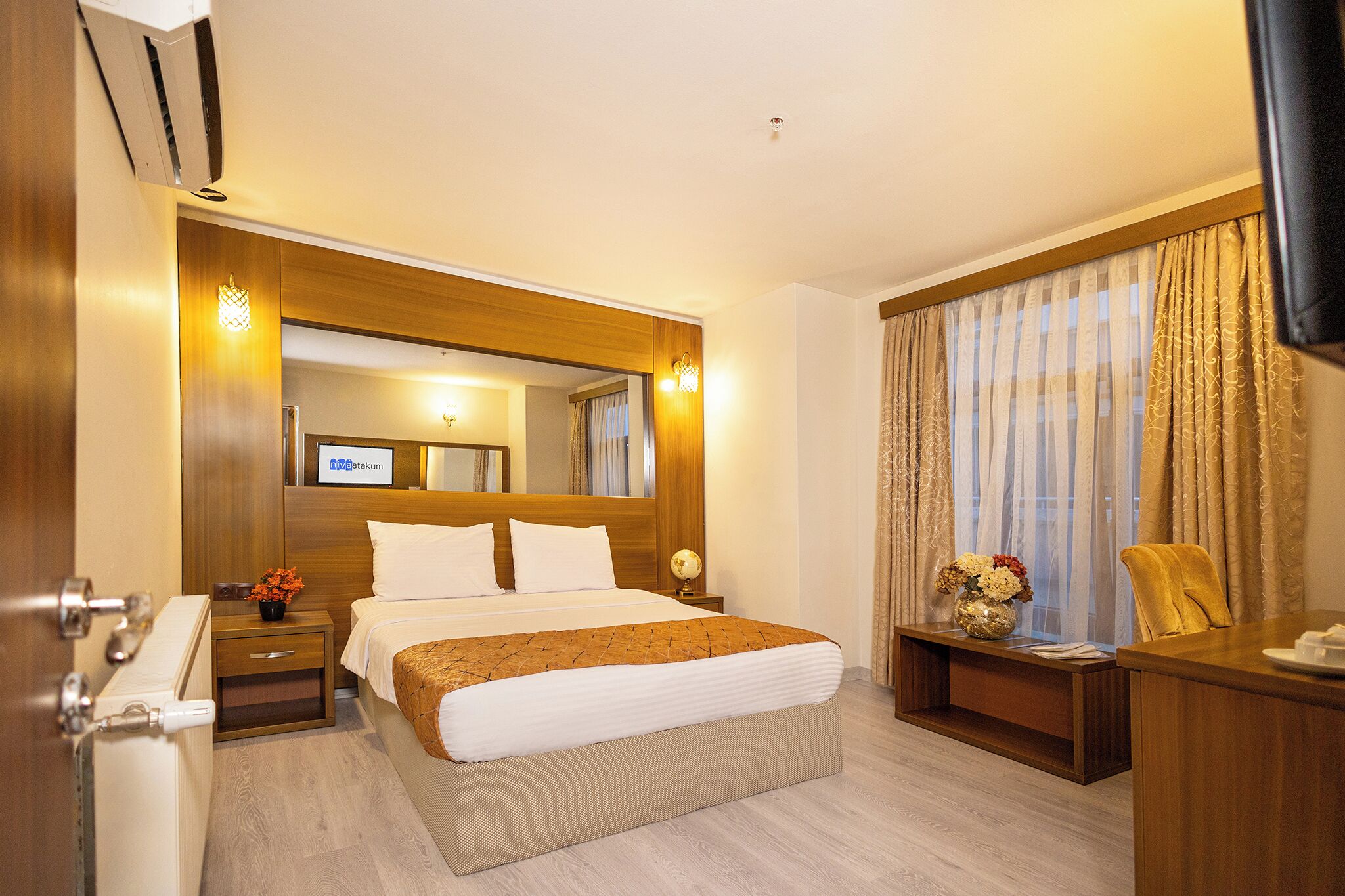 A hotel room with a large bed, wooden headboard, a desk, a chair, a TV, and a window with curtains.