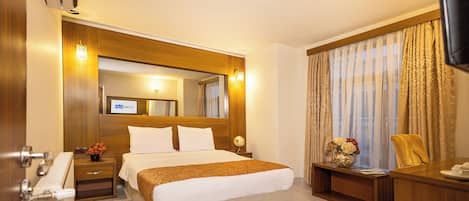 Habitación de hotel con una cama grande, cabecera de madera, un escritorio, una silla, un televisor y una ventana con cortinas.