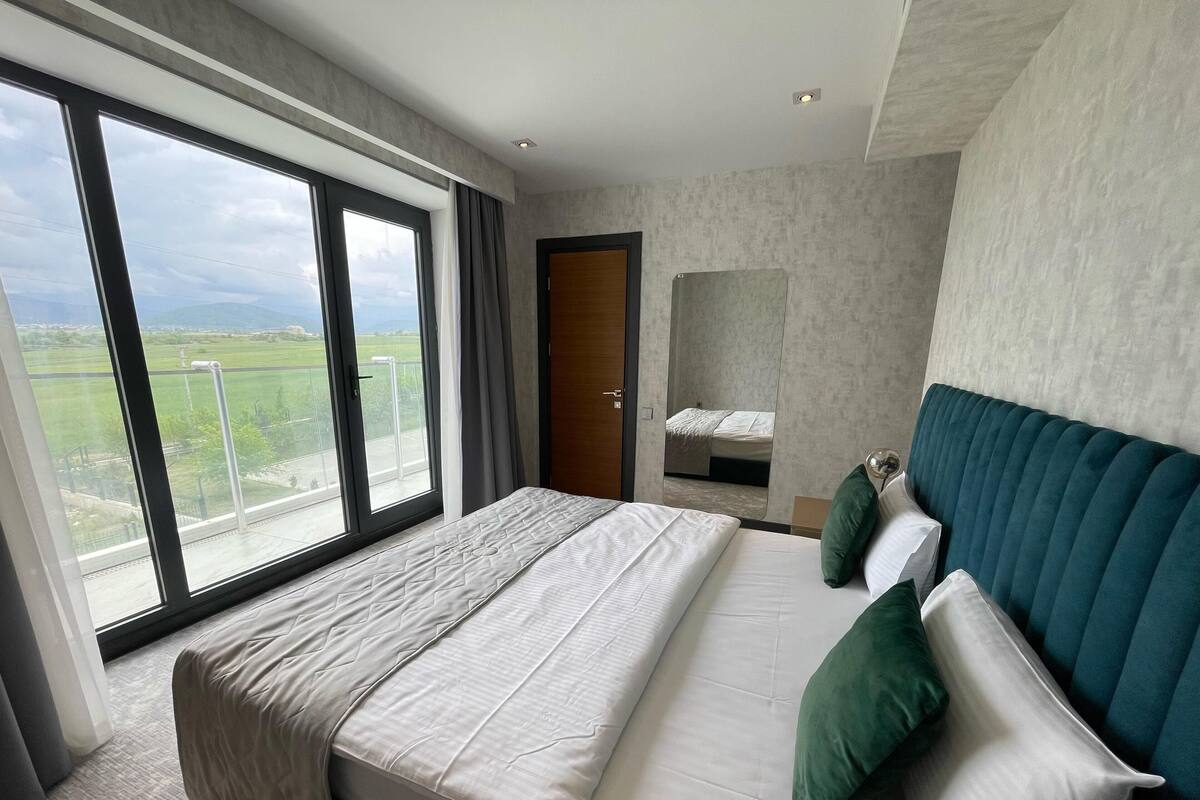 A modern hotel room with a large bed, a view of a green landscape through floor-to-ceiling windows, and a balcony.