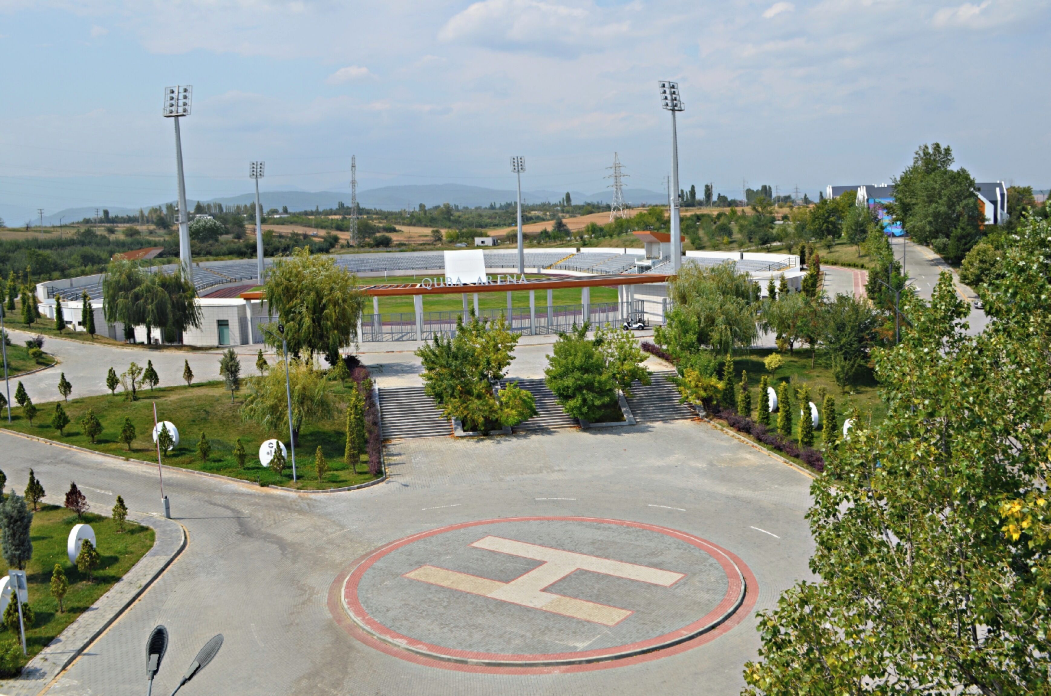 Un stade avec un grand marquage en forme de H sur le terrain, entouré d’arbres et doté d’un parking.