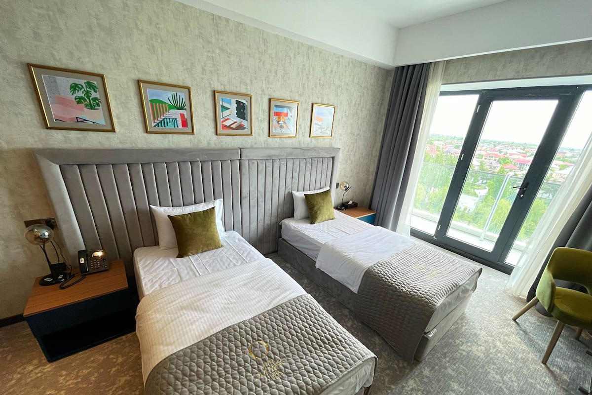 A hotel room with two beds, a large window with a city view, and artwork on the wall.