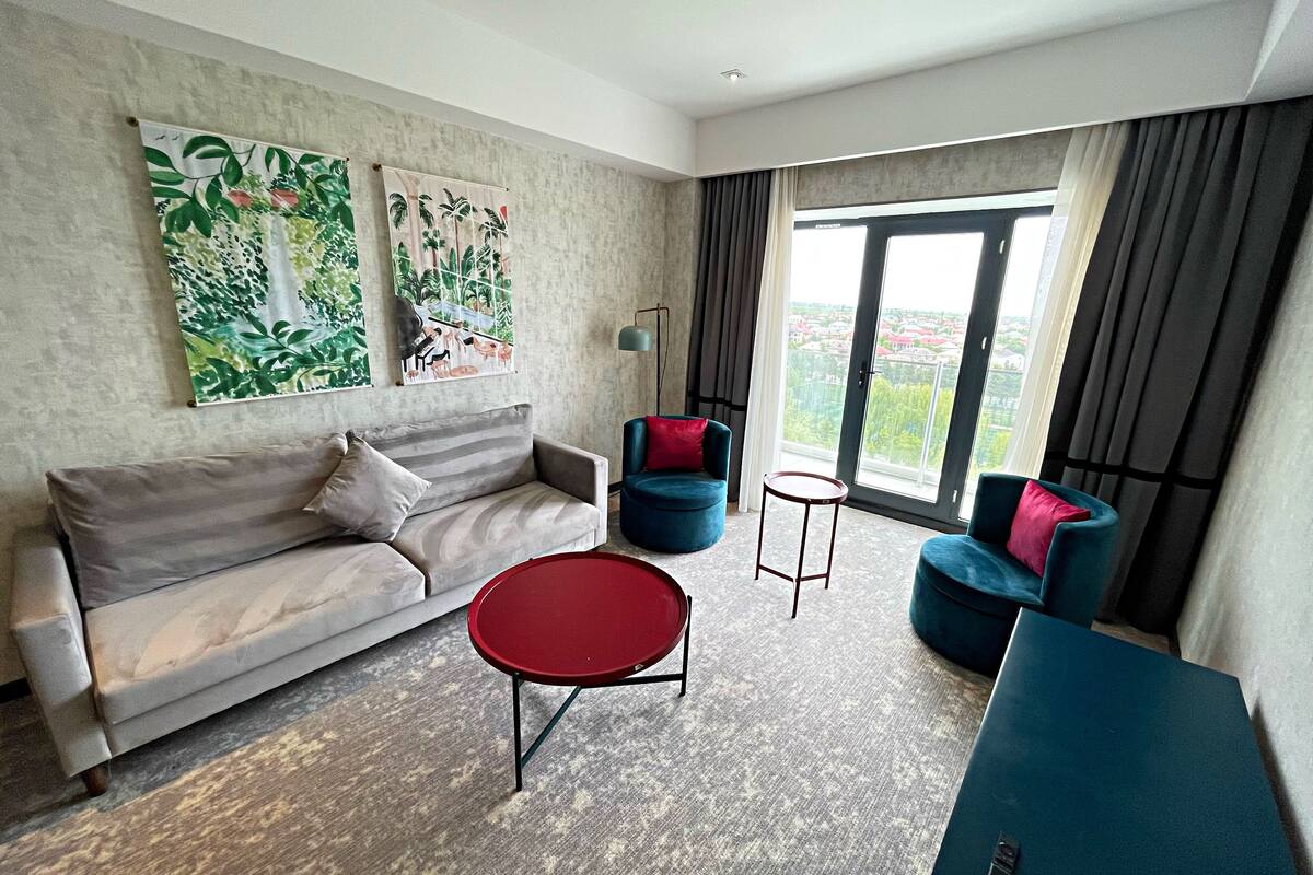 A modern living room with a grey sofa, red coffee table, and blue armchairs. There are two large paintings on the wall and a view of the city through the window.