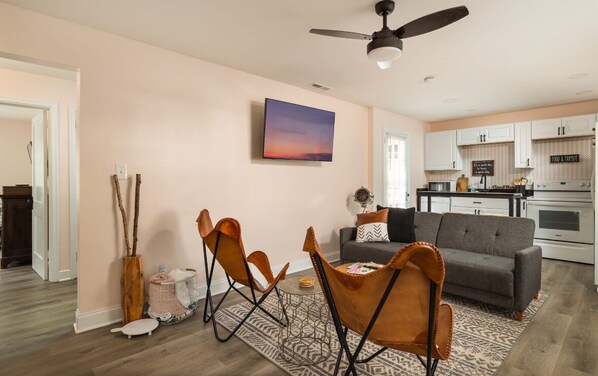 Una sala de estar moderna con un sofá gris, una mesa de centro y un comedor con sillas. Se ve una cocina al fondo con armarios blancos y un letrero que dice "Food & Friends".