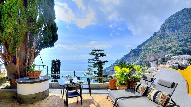 Une terrasse avec une table et des chaises, des plantes en pot, et une vue sur la mer et les montagnes.