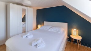Un dormitorio con una cama grande, ropa de cama blanca, un armario, una lámpara de noche y un techo inclinado.
