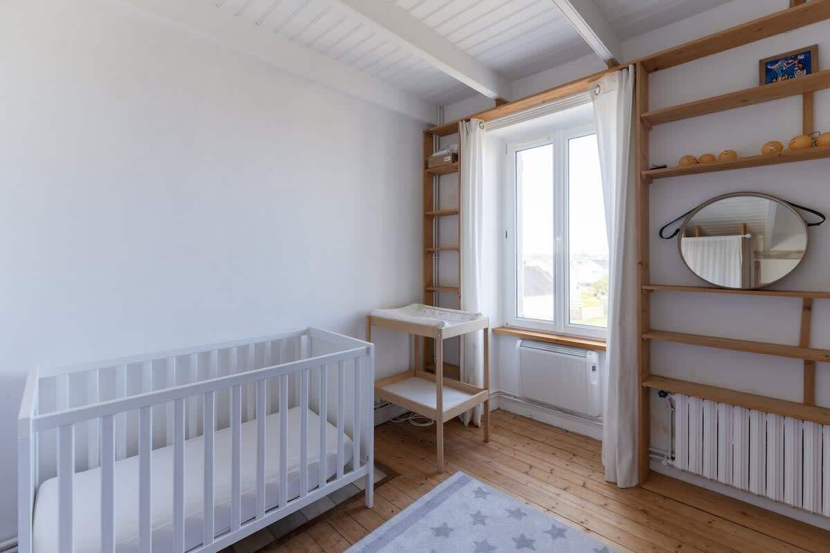 Ein Babyzimmer mit weißem Kinderbett, kleinem Tisch, einem Fenster mit Vorhängen und einem Regal mit Babyartikeln.