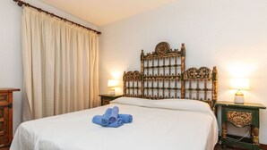 Un lit bien fait, recouvert d’une courtepointe blanche et agrémenté de serviettes bleues, une tête de lit en bois ornée de motifs complexes, deux lampes de chevet et une table de chevet en bois sur laquelle est posé un objet décoratif.