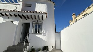 A white building with a tiled roof, a window with metal bars, and a door. There are steps leading up to the entrance, and three potted plants are placed near the door.