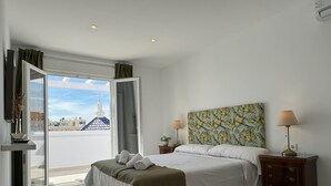 A modern hotel room with a large bed, two bedside tables, a view of a cityscape through an open door, and a ceiling with recessed lighting.