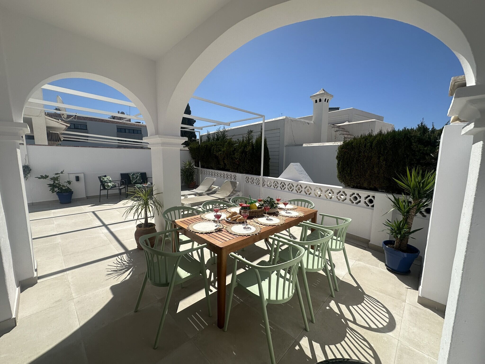 Une terrasse extérieure avec une table en bois dressée pour le repas, entourée de chaises vertes, sous des arches blanches.