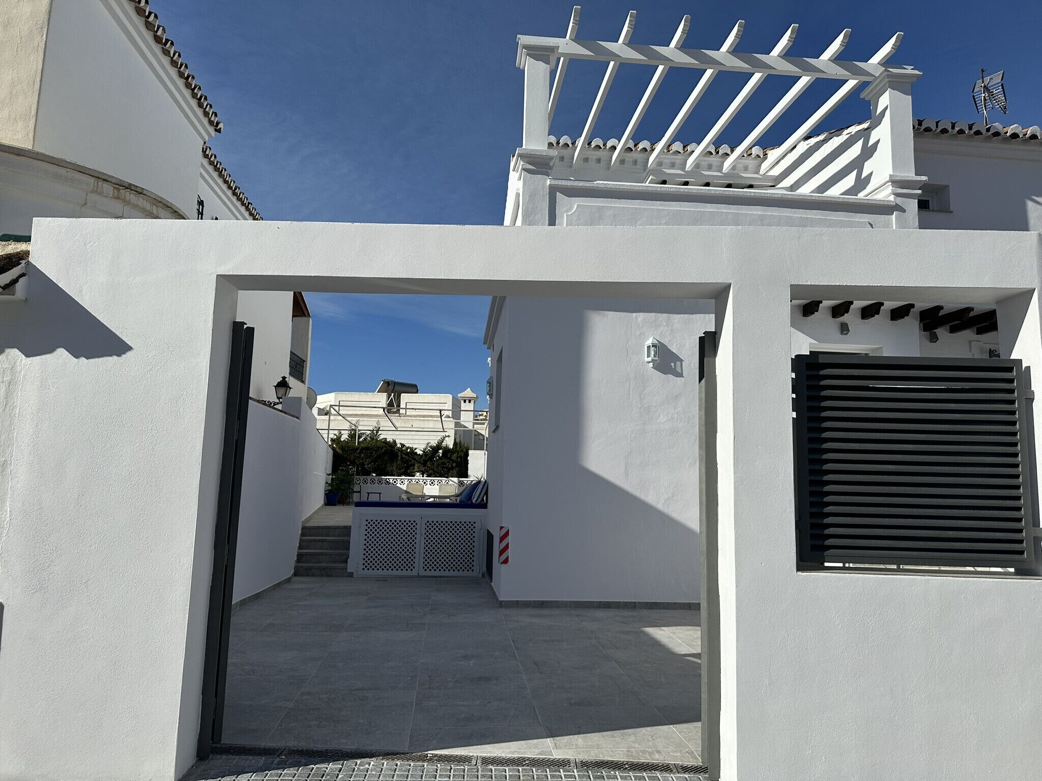 Un edificio blanco con un espacio exterior techado, una entrada protegida por una reja y vistas a un edificio distante.