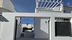 Un bâtiment blanc doté d’un espace extérieur couvert, d’une entrée fermée par un portail et d’où l’on peut apercevoir un autre bâtiment situé au loin.
