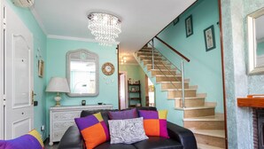 A living room with a black leather sofa, colorful cushions, a white coffee table with pastries, a chandelier, and a staircase.