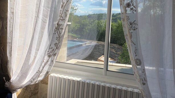 A room with a window featuring floral curtains, a radiator below, and a view of greenery and a swimming pool outside.