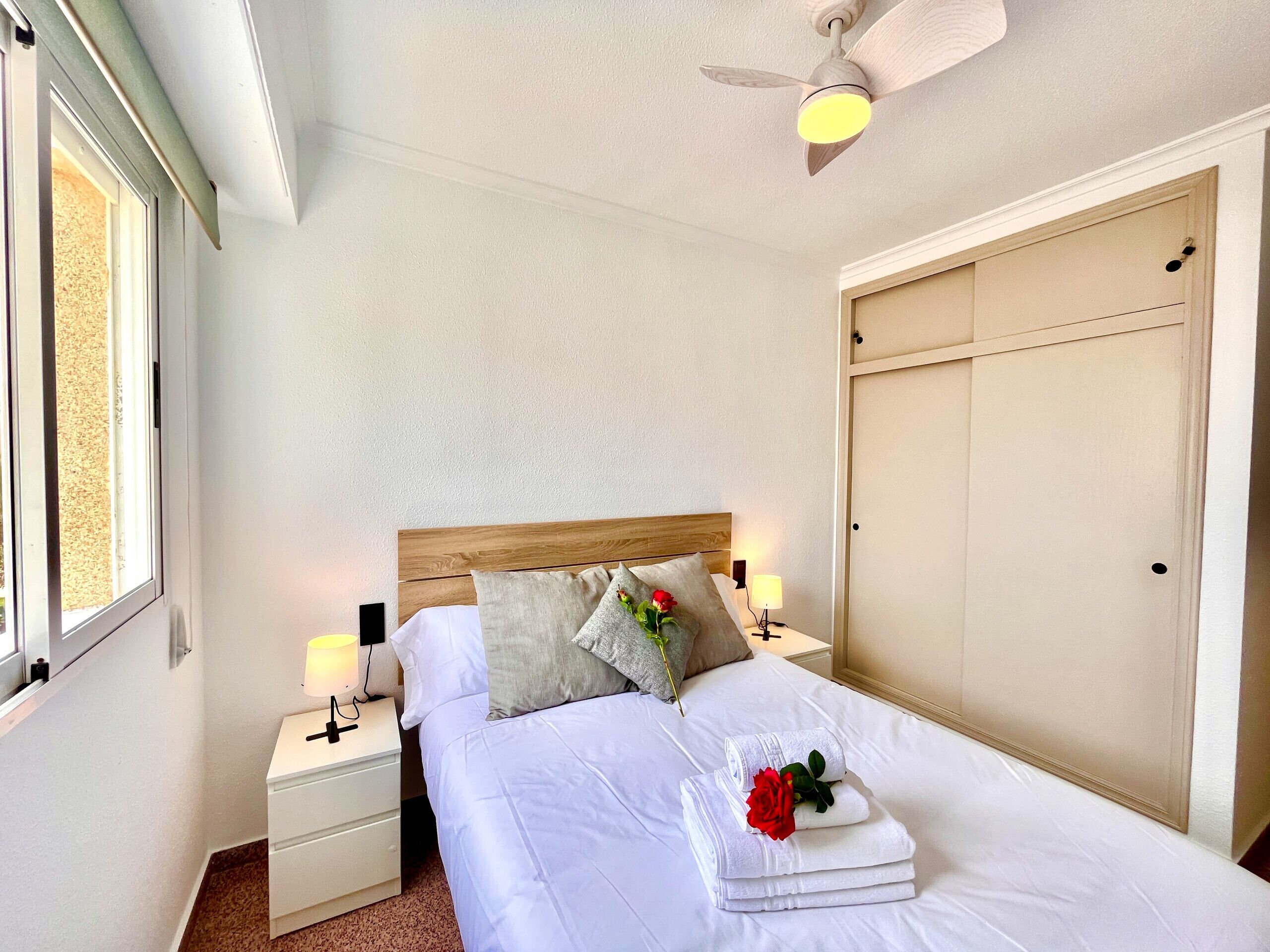 A neatly made bed with white linens, a wooden headboard, and a nightstand with a lamp. There is a window on the left side, a door on the right, and a ceiling fan above.