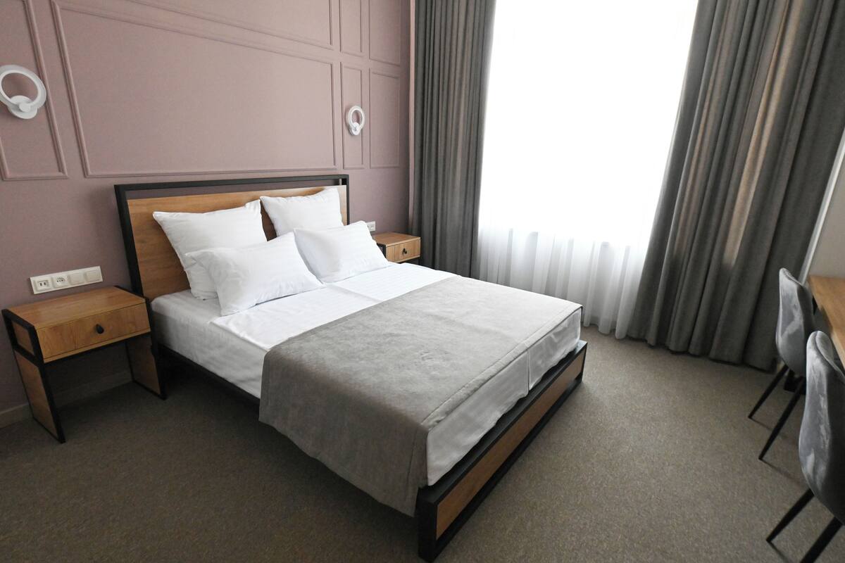 Un lit bien fait, avec du linge de lit blanc, une tête de lit en bois et une couvre-lit grise. Il y a deux tables de chevet avec des tiroirs, une de chaque côté du lit. La pièce est décorée dans des tons neutres, avec un rideau gris à la fenêtre.