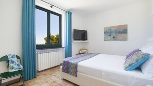 Un dormitorio con cama, televisión, un cuadro, una silla y una ventana con cortinas.
