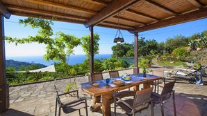 Un patio exterior con mesa y sillas de madera, con vistas a un paisaje pintoresco con árboles y el mar.