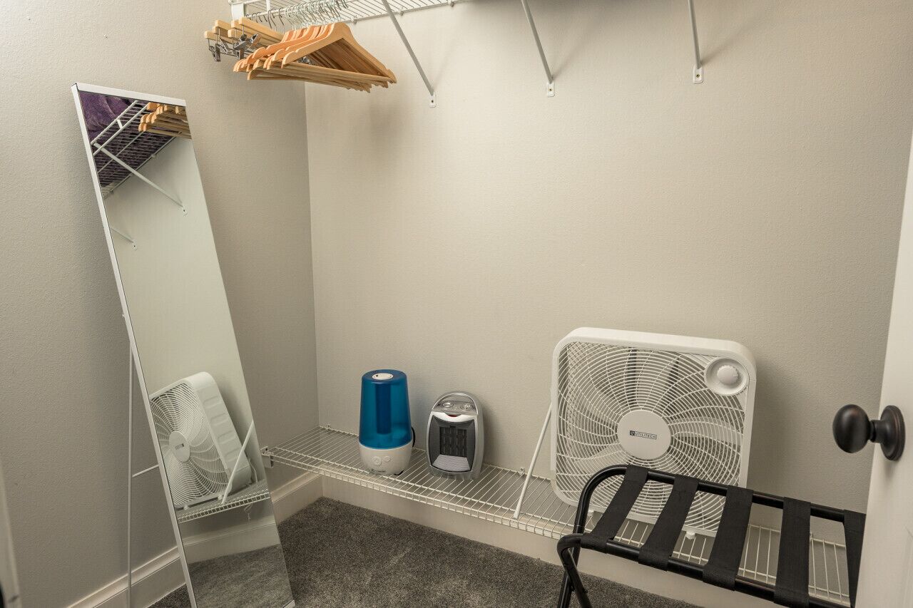 Une salle de bain avec un miroir, un ventilateur blanc, un chauffe-eau bleu et un radiateur électrique blanc posé sur une étagère.