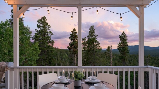 Un espacio de comedor al aire libre con techo, con una mesa dispuesta para cuatro personas, rodeada de sillas blancas, con vistas a un paisaje boscoso y montañas a lo lejos.
