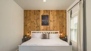 Un dormitorio con pared de paneles de madera, una cama blanca con cabecera, mesitas de noche con lámparas y un ventanal con cortinas.