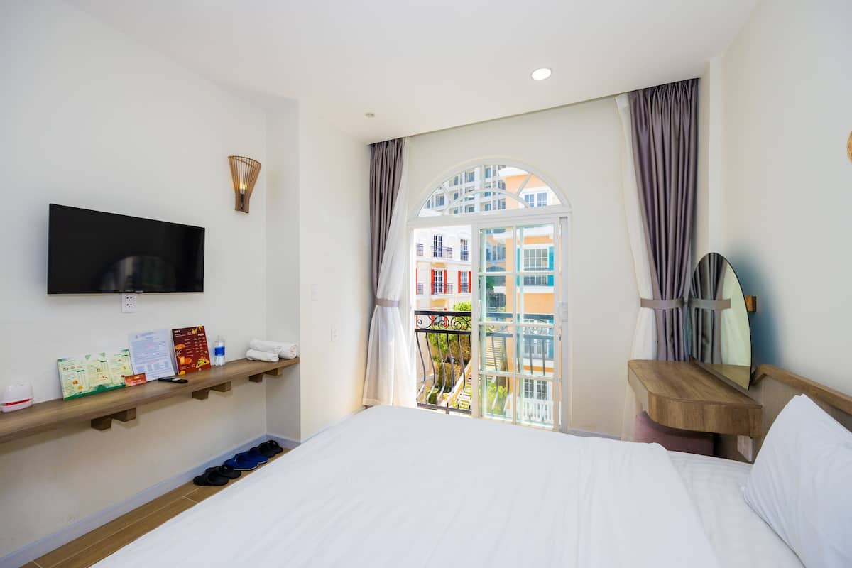 A hotel room with a bed, a TV mounted on the wall, a wooden shelf with toiletries, a window with a view of buildings, and a mirror on the wall.